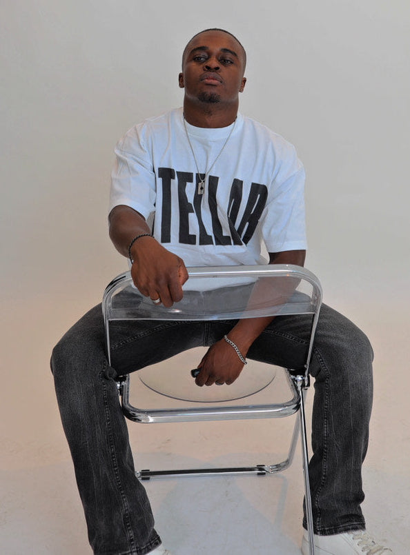 Man sitting on a clear chair wearing a white t-shirt with 'TELLLO' printed on it against a plain background.